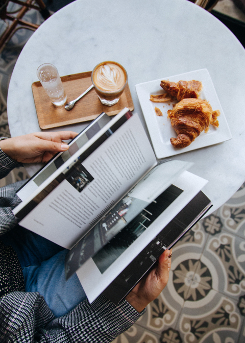 Person reading coffee table book at round table with water, coffee, and croissant.