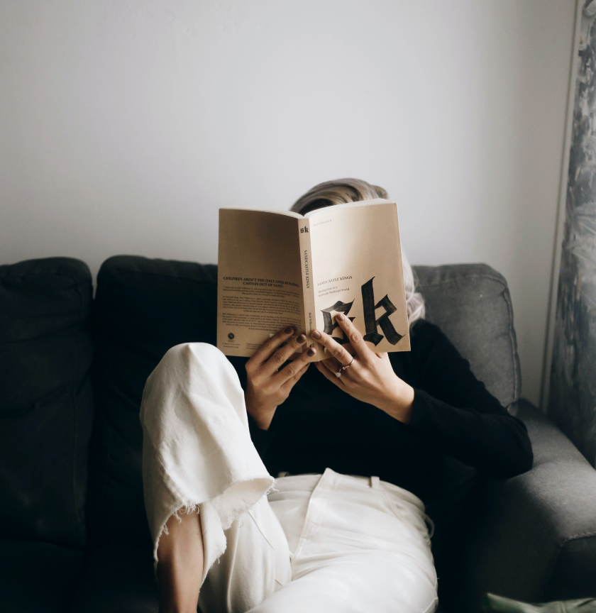 Person in white pants and black shirt sitting on couch reading from book.